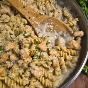 Creamy Garlic Parmesan Chicken Pasta 3 Close-up overhead view of Creamy Garlic Parmesan Chicken Pasta in a metal pan with a wooden spoon, garnished with fresh herbs and Parmesan.