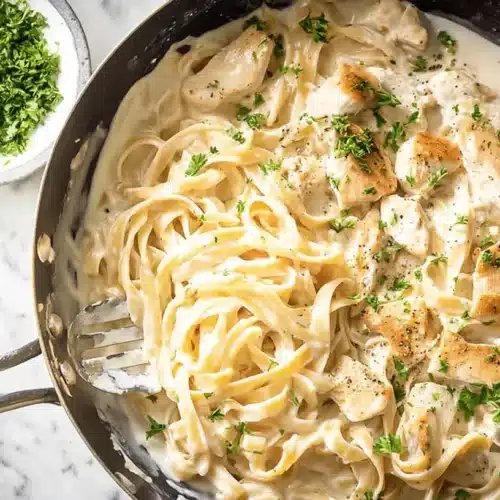 Overhead view of a delicious creamy chicken alfredo pasta dish garnished with fresh parsley in a dark pan on a marble surface.
