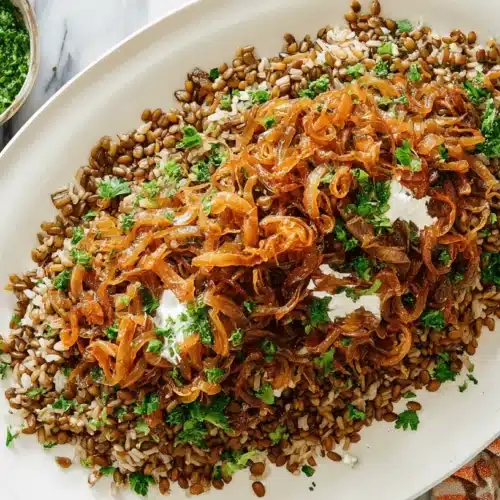 Hearty Mujadara Recipe 1 A hearty platter of Mujadara (Lentils and Rice with Caramelized Onions) garnished with fresh herbs and creamy yogurt, served alongside green herb sauce and a creamy dip on a marble counter.