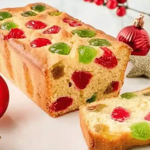 A festive Newfoundland Cherry Cake, golden-brown with red and green candied cherries, presented on a marble surface with Christmas ornaments and a sliced piece revealing its moist, pale yellow interior.