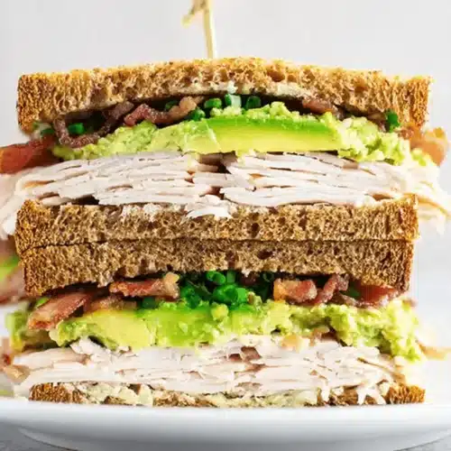 The Ultimate Turkey and Avocado Sandwich Recipe 1 A tall, triple-decker turkey and avocado sandwich with bacon and chives, served on toasted whole wheat bread.