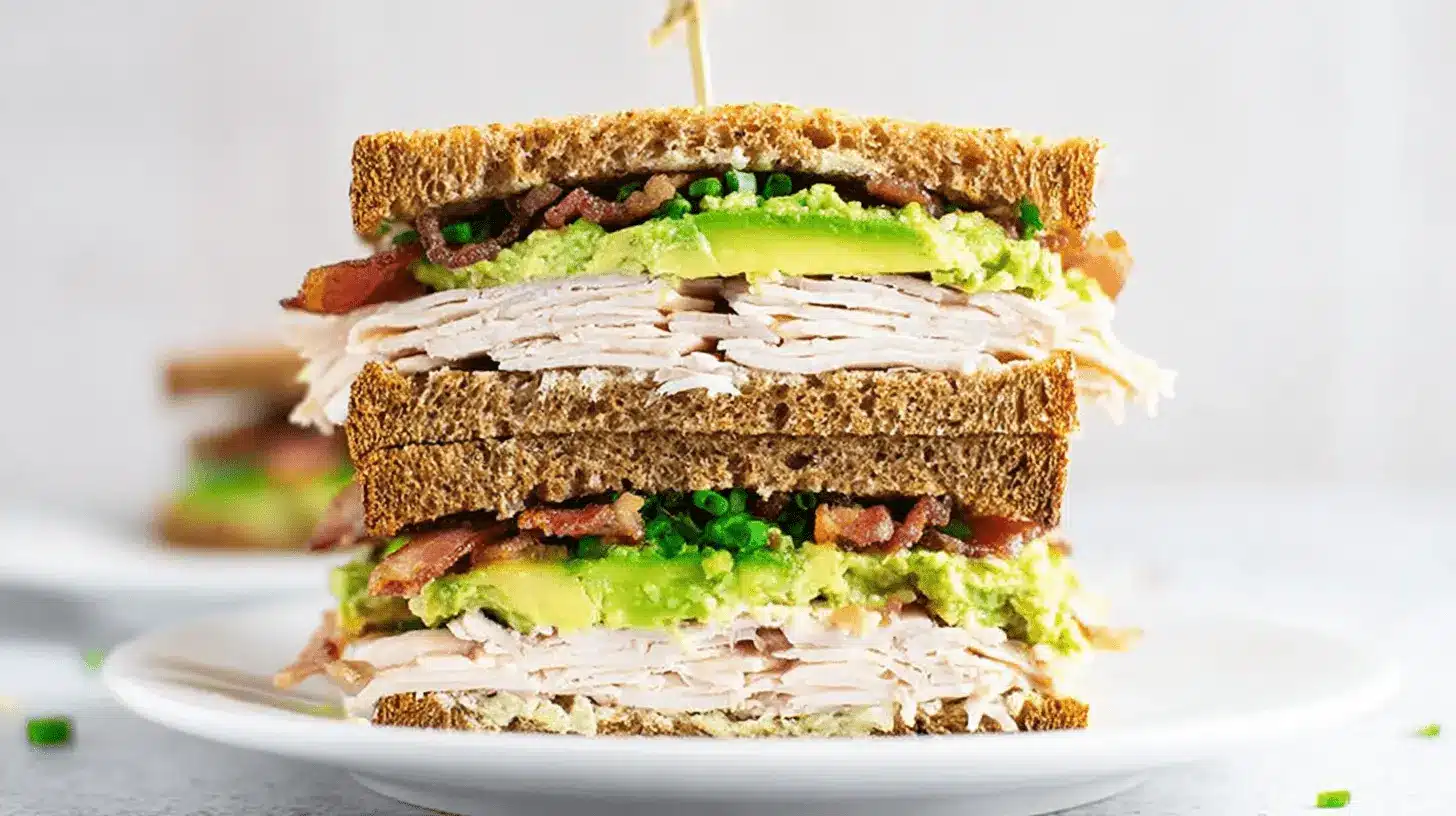 A tall, triple-decker turkey and avocado sandwich with bacon and chives, served on toasted whole wheat bread.